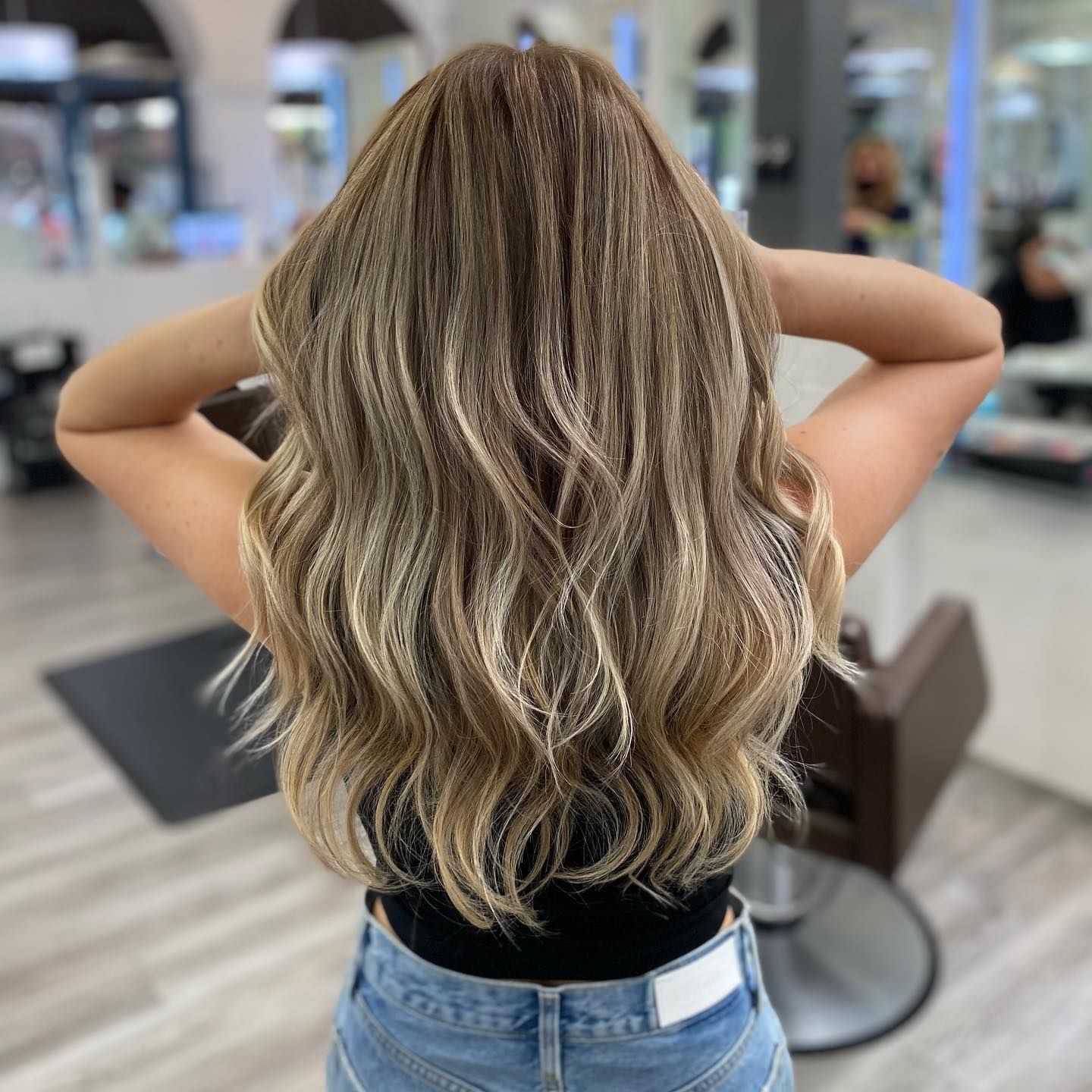 Woman with long, wavy blonde hair in a salon setting, viewed from the back.